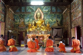 A group of seven monks in orange robes are kneeling in worship inside a richly decorated temple. The room features an ornate altar with a large golden Buddha statue surrounded by smaller statues. The walls are adorned with colorful murals depicting scenes of Buddhist significance, and delicate architectural details are visible in the background.