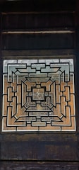 A geometric, symmetrical pattern formed by interlocking lines creates a complex design within a rectangular frame. The central architectural element is set against a wooden backdrop, displaying intricate craftsmanship.