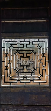 A geometric, symmetrical pattern formed by interlocking lines creates a complex design within a rectangular frame. The central architectural element is set against a wooden backdrop, displaying intricate craftsmanship.