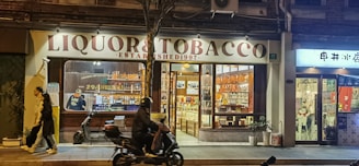 A nighttime street scene features a store named 'Liquor & Tobacco' with a well-lit interior displaying bottles and products on shelves. A person on a motorbike rides past, while another pedestrian walks by on the sidewalk. The atmosphere is vibrant with artificial lighting from the store windows.