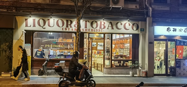 A nighttime street scene features a store named 'Liquor & Tobacco' with a well-lit interior displaying bottles and products on shelves. A person on a motorbike rides past, while another pedestrian walks by on the sidewalk. The atmosphere is vibrant with artificial lighting from the store windows.