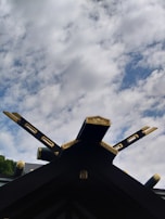A traditional roof structure with ornate golden decorations and intersecting beams extends upward against a backdrop of a partly cloudy sky. The beams are intricately carved and feature gold accents, adding a touch of elegance to the architectural design.