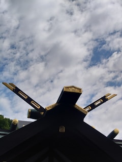 A traditional roof structure with ornate golden decorations and intersecting beams extends upward against a backdrop of a partly cloudy sky. The beams are intricately carved and feature gold accents, adding a touch of elegance to the architectural design.