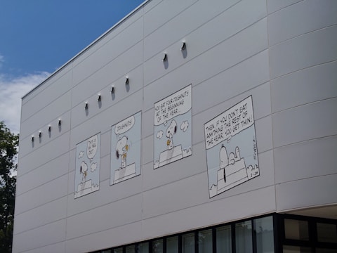 A building facade features a series of four comic strip panels depicting a cartoon character talking about a new diet involving doughnuts. The panels are aligned horizontally across the exterior wall. The artwork is in black and white, with the comic character prominently featured.