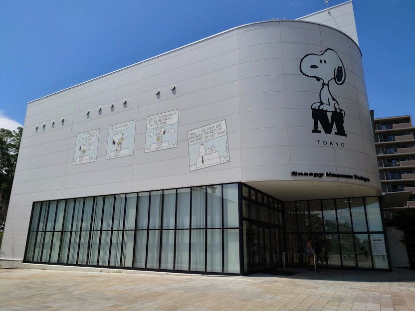 The building exterior features a large cartoon design with characters from the Peanuts comic strip, prominently displaying Snoopy. The name 'Snoopy Museum Tokyo' is indicated on the building. The structure is modern, with large glass windows on the ground floor and cartoon panels adorning the upper walls.