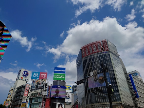 A bustling urban scene with tall buildings adorned with colorful advertisements and billboards against a partly cloudy blue sky. Pedestrian traffic lights and electronic screens featuring various images and text are visible. The area is vibrant and modern, suggesting a commercial or shopping district.