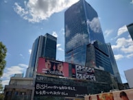 A cityscape featuring tall modern skyscrapers with reflective glass facades under a partly cloudy sky. Prominent advertisements are visible on the buildings, including one for SoftBank and another for a Netflix show.