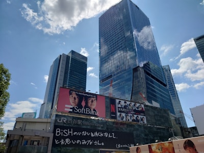 A cityscape featuring tall modern skyscrapers with reflective glass facades under a partly cloudy sky. Prominent advertisements are visible on the buildings, including one for SoftBank and another for a Netflix show.