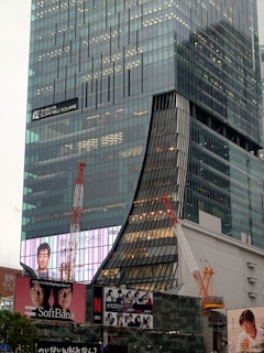 A modern high-rise building with glass paneling dominates the scene. The structure features a unique curved architectural design with construction cranes positioned nearby. Large digital billboards display advertisements, with one featuring a person smiling and others showcasing groups of people. The signage indicates it is Shibuya Scramble Square.