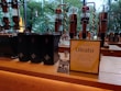 A modern café counter featuring a display of coffee-making equipment and containers. There are several tall, transparent glass apparatuses with copper spiral tubes, and black containers with star logos. A standing sign and a small promotional card introduce a new product called Oleato. Large windows in the background offer a view of green trees outside.