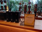 A modern café counter featuring a display of coffee-making equipment and containers. There are several tall, transparent glass apparatuses with copper spiral tubes, and black containers with star logos. A standing sign and a small promotional card introduce a new product called Oleato. Large windows in the background offer a view of green trees outside.