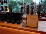 A modern café counter featuring a display of coffee-making equipment and containers. There are several tall, transparent glass apparatuses with copper spiral tubes, and black containers with star logos. A standing sign and a small promotional card introduce a new product called Oleato. Large windows in the background offer a view of green trees outside.