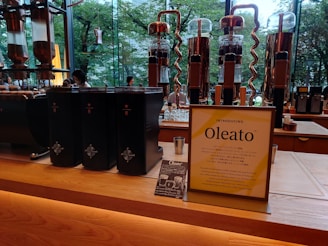 A modern café counter featuring a display of coffee-making equipment and containers. There are several tall, transparent glass apparatuses with copper spiral tubes, and black containers with star logos. A standing sign and a small promotional card introduce a new product called Oleato. Large windows in the background offer a view of green trees outside.