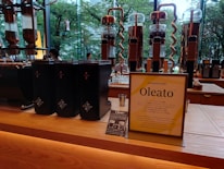 A modern café counter featuring a display of coffee-making equipment and containers. There are several tall, transparent glass apparatuses with copper spiral tubes, and black containers with star logos. A standing sign and a small promotional card introduce a new product called Oleato. Large windows in the background offer a view of green trees outside.