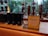 A modern café counter featuring a display of coffee-making equipment and containers. There are several tall, transparent glass apparatuses with copper spiral tubes, and black containers with star logos. A standing sign and a small promotional card introduce a new product called Oleato. Large windows in the background offer a view of green trees outside.