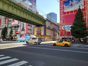 A bustling city intersection features colorful buildings with various advertisements and billboards. A yellow sports car and a white van are visible on the road. There is a large overhead railway bridge, and numerous people are walking along the sidewalks. One building has a large tree next to it.