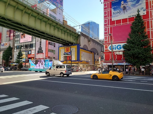 A bustling city intersection features colorful buildings with various advertisements and billboards. A yellow sports car and a white van are visible on the road. There is a large overhead railway bridge, and numerous people are walking along the sidewalks. One building has a large tree next to it.