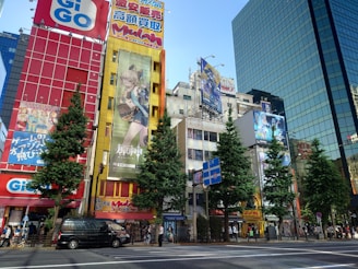 A bustling urban scene with tall buildings adorned with colorful advertisements and banners. The foreground features a busy street with pedestrians and vehicles, while several trees line the sidewalk. The bright colors and bold designs of the ads create a vibrant city atmosphere.