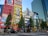 A bustling urban scene with tall buildings adorned with colorful advertisements and banners. The foreground features a busy street with pedestrians and vehicles, while several trees line the sidewalk. The bright colors and bold designs of the ads create a vibrant city atmosphere.