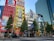 A bustling urban scene with tall buildings adorned with colorful advertisements and banners. The foreground features a busy street with pedestrians and vehicles, while several trees line the sidewalk. The bright colors and bold designs of the ads create a vibrant city atmosphere.