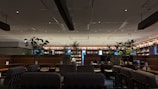 A modern, spacious indoor setting with contemporary seating arrangements. There are several plants and greenery placed throughout the room, contributing to a calming atmosphere. Shelving units are visible in the background, likely holding books or decorative items. The space also has multiple screens on a table, possibly indicating a shared workspace or study area. The lighting is soft and subtle, enhancing the cozy and comfortable environment.