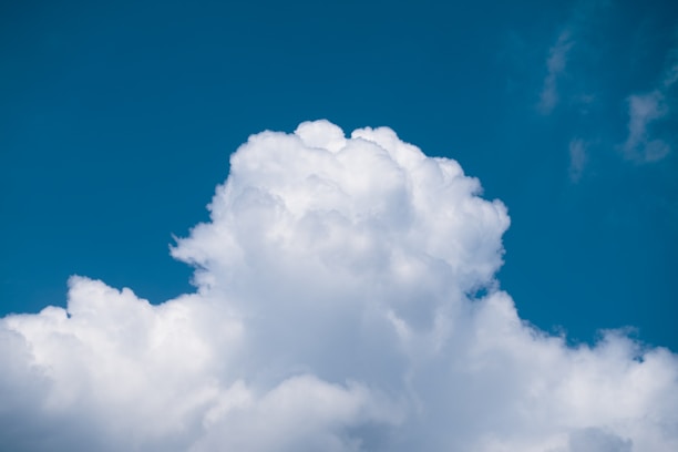 A large, fluffy cumulus cloud fills the foreground, contrasting sharply with a clear, vibrant blue sky in the background. The cloud appears soft and voluminous, with its bright white color enhanced by sunlight.