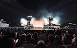 A large crowd gathers in a stadium, facing toward a brightly lit stage with multiple spotlights and large screens. Two camera rigs are positioned prominently above the crowd, capturing the event. The atmosphere suggests a concert or a large public gathering.