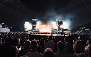 A large crowd gathers in a stadium, facing toward a brightly lit stage with multiple spotlights and large screens. Two camera rigs are positioned prominently above the crowd, capturing the event. The atmosphere suggests a concert or a large public gathering.