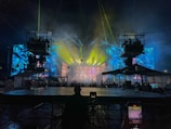 A vibrant concert stage with elaborate lighting effects. Bright yellow, blue, and green lights create an electrifying atmosphere. Large screens display colorful abstract visuals. Scaffolding surrounds the stage, indicating a large production setup. An audience is visible, adding to the lively atmosphere.