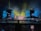 A vibrant concert stage with elaborate lighting effects. Bright yellow, blue, and green lights create an electrifying atmosphere. Large screens display colorful abstract visuals. Scaffolding surrounds the stage, indicating a large production setup. An audience is visible, adding to the lively atmosphere.