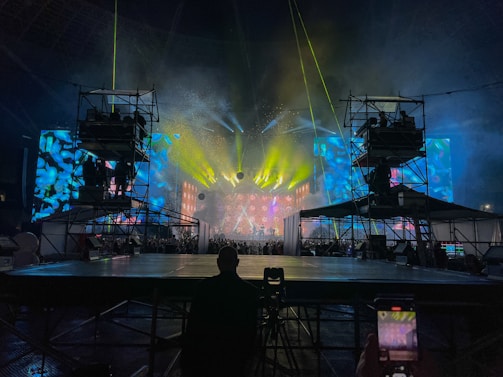 A vibrant concert stage with elaborate lighting effects. Bright yellow, blue, and green lights create an electrifying atmosphere. Large screens display colorful abstract visuals. Scaffolding surrounds the stage, indicating a large production setup. An audience is visible, adding to the lively atmosphere.