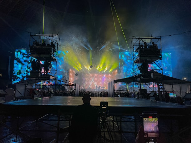 A vibrant concert stage with elaborate lighting effects. Bright yellow, blue, and green lights create an electrifying atmosphere. Large screens display colorful abstract visuals. Scaffolding surrounds the stage, indicating a large production setup. An audience is visible, adding to the lively atmosphere.