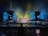 A vibrant concert stage with elaborate lighting effects. Bright yellow, blue, and green lights create an electrifying atmosphere. Large screens display colorful abstract visuals. Scaffolding surrounds the stage, indicating a large production setup. An audience is visible, adding to the lively atmosphere.