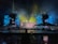 A vibrant concert stage with elaborate lighting effects. Bright yellow, blue, and green lights create an electrifying atmosphere. Large screens display colorful abstract visuals. Scaffolding surrounds the stage, indicating a large production setup. An audience is visible, adding to the lively atmosphere.