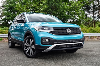 A turquoise SUV is parked on a paved road with lush green trees and a metallic guardrail in the background. The vehicle has pronounced black wheel arches and a distinctive front grille with the Volkswagen logo prominently displayed.