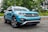 A turquoise SUV is parked on a paved road with lush green trees and a metallic guardrail in the background. The vehicle has pronounced black wheel arches and a distinctive front grille with the Volkswagen logo prominently displayed.