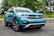 A turquoise SUV is parked on a paved road with lush green trees and a metallic guardrail in the background. The vehicle has pronounced black wheel arches and a distinctive front grille with the Volkswagen logo prominently displayed.