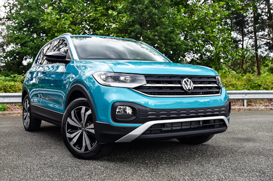 A turquoise SUV is parked on a paved road with lush green trees and a metallic guardrail in the background. The vehicle has pronounced black wheel arches and a distinctive front grille with the Volkswagen logo prominently displayed.