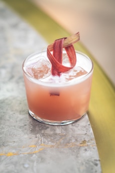 A cocktail in a transparent glass featuring a light pink hue, topped with foam and ice cubes. A decorative garnish made of twisted rhubarb sits on top. Positioned on a marble surface with warm tones.
