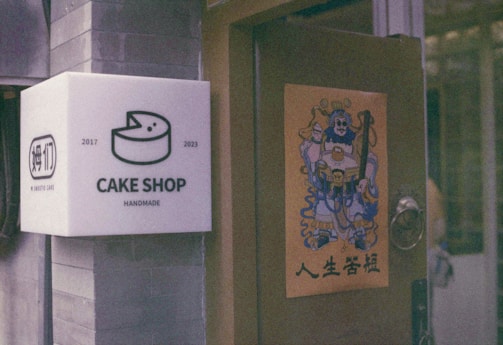 A cake shop sign mounted on a wall featuring an illustration of a cake, along with the years 2017 and 2023 indicating its establishment. Adjacent, a door displays a poster with a colorful, traditional Chinese warrior design and Chinese characters beneath it.