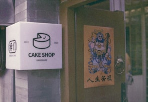 A cake shop sign mounted on a wall featuring an illustration of a cake, along with the years 2017 and 2023 indicating its establishment. Adjacent, a door displays a poster with a colorful, traditional Chinese warrior design and Chinese characters beneath it.
