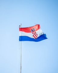 A flag with three horizontal stripes of red, white, and blue is waving against a clear blue sky. The coat of arms is prominently centered on the white stripe.