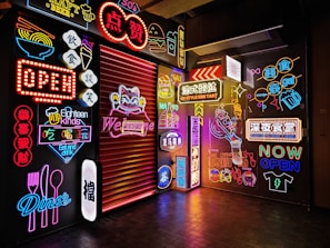 A vibrant array of colorful neon signs featuring various designs and text covers the walls. Some signs depict food-related images such as bowls, forks, and spoons, while others display open and welcome messages. A large glowing cat with a welcoming gesture is prominently featured alongside numerous Chinese characters and decorative graphics.