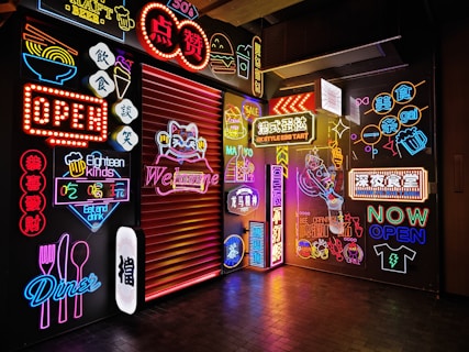 A vibrant array of colorful neon signs featuring various designs and text covers the walls. Some signs depict food-related images such as bowls, forks, and spoons, while others display open and welcome messages. A large glowing cat with a welcoming gesture is prominently featured alongside numerous Chinese characters and decorative graphics.