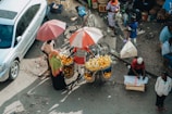 Aerial view of a busy street market where a vendor with a bicycle is selling oranges from large baskets under umbrellas. Several people are engaged in various activities around the market, and a car is parked on the left side of the scene.