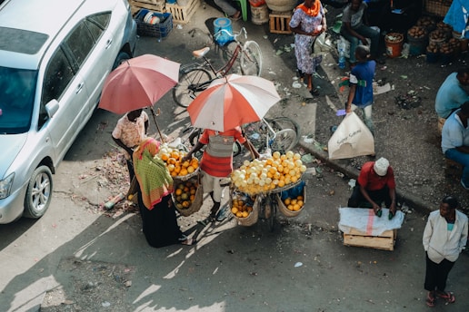 Aerial view of a busy street market where a vendor with a bicycle is selling oranges from large baskets under umbrellas. Several people are engaged in various activities around the market, and a car is parked on the left side of the scene.