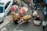 Aerial view of a busy street market where a vendor with a bicycle is selling oranges from large baskets under umbrellas. Several people are engaged in various activities around the market, and a car is parked on the left side of the scene.