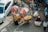 Aerial view of a busy street market where a vendor with a bicycle is selling oranges from large baskets under umbrellas. Several people are engaged in various activities around the market, and a car is parked on the left side of the scene.