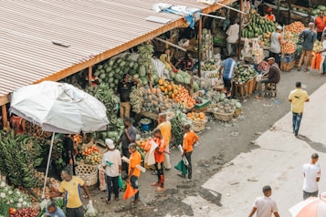 A bustling outdoor market scene with multiple stalls displaying a wide variety of fresh fruits and vegetables. Several people are shopping, browsing, and interacting with vendors. An array of bananas, pineapples, watermelons, and other colorful produce are arranged in baskets and crates. The market is covered by a simple metal roof, and one large umbrella provides shade for part of the market. The ground is littered with small debris, adding an authentic, lived-in feel to the environment.