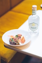 A white plate on a table holds a gourmet dish featuring colorful garnishes, grilled vegetables, and a delicate, lacy edible decoration. Next to the plate, there is a clear glass bottle with a cork stopper and a label displaying the word 'Anaë'. The background includes a soft, warm-toned cushioned seat.